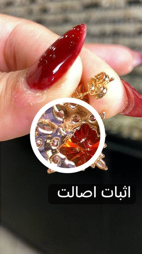 تصویر نمای نزدیک گوشواره سوزنی ژوپینگ اورجینال مدل سولار، با نگین مرکزی قرمز و جزئیات ساخت و منافذ مخراجکاری در نمای پشت، همراه با نمایش اثبات اصالت محصول