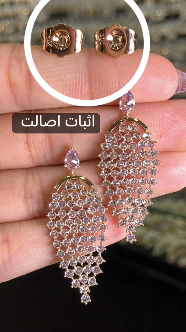 تصویر جفت گوشواره ژوپینگ اورجینال روی دست، با طراحی بال فرشته، نگینهای اتمی سفید و شفاف با کاشت معکوس، آبکاری طلایی و نمای نزدیک پشتهای سوزنی برای نمایش جزئیات و اثبات اصالت ژوپینگ