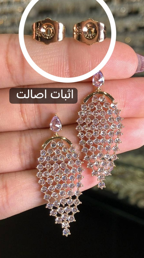 تصویر جفت گوشواره ژوپینگ اورجینال روی دست، با طراحی بال فرشته، نگینهای اتمی سفید و شفاف با کاشت معکوس، آبکاری طلایی و نمای نزدیک پشتهای سوزنی برای نمایش جزئیات و اثبات اصالت ژوپینگ